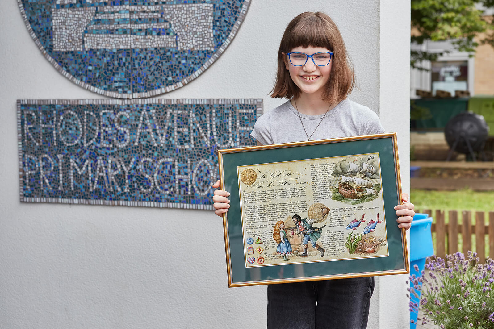 A child stands in front of their school sign holding an illustrated framed copy of their winning short story